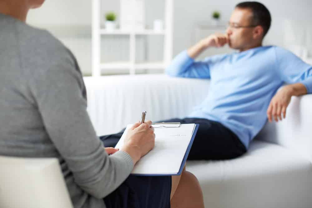 Thoughtful man on couch looking away, therapist in foreground, cropped